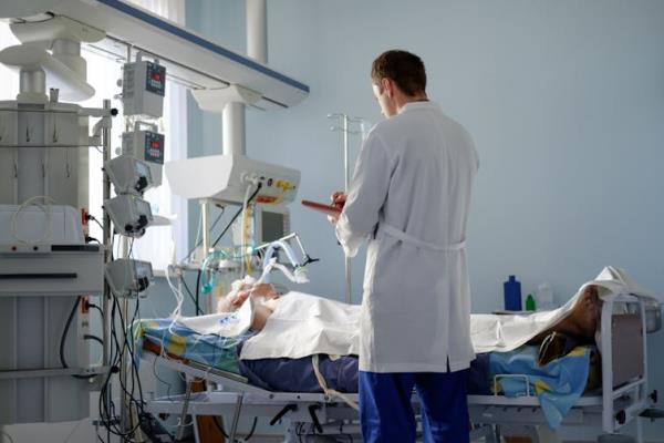 A doctor with a clipboard stands at a patient's bedside in an ICU.