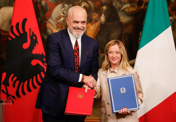 Edi Rama and Giorgia Meloni shake hands while holding diplomatic folders, standing in front of Albanian and Italian flags