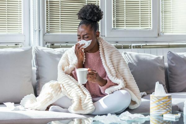 A sick woman sneezes into a tissue while sitting on her couch.