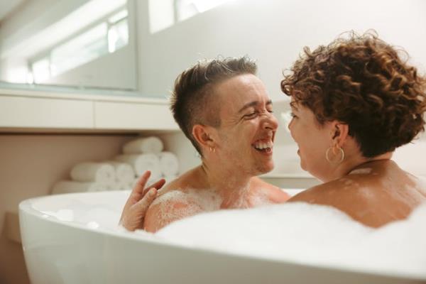 A queer couple laughing together in a bath.